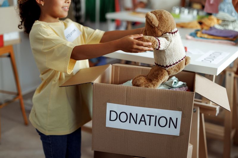Young girl volunteering by gently placing a teddy bear into a donation box. Surrounded by other donated items, she represents kindness, generosity, and youth involvement in community service. Ideal for themes of charity, giving, and child volunteerism