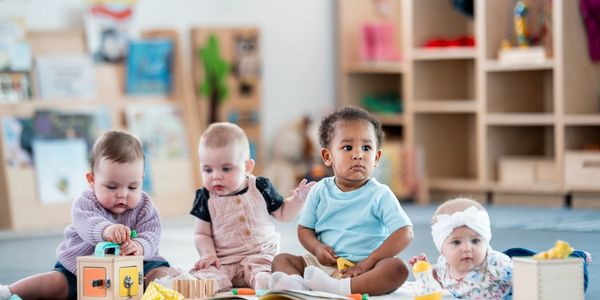 Four babies playing together on a mat with toys in a bright playroom.