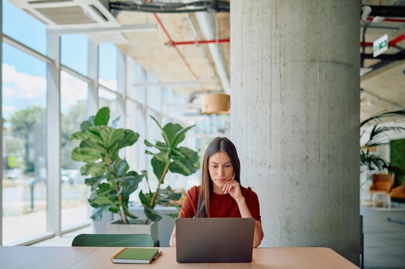 Young businesswoman concentrating while using laptop at desk in bright, modern office space with large windows and plants, enjoying the natural light and greenery