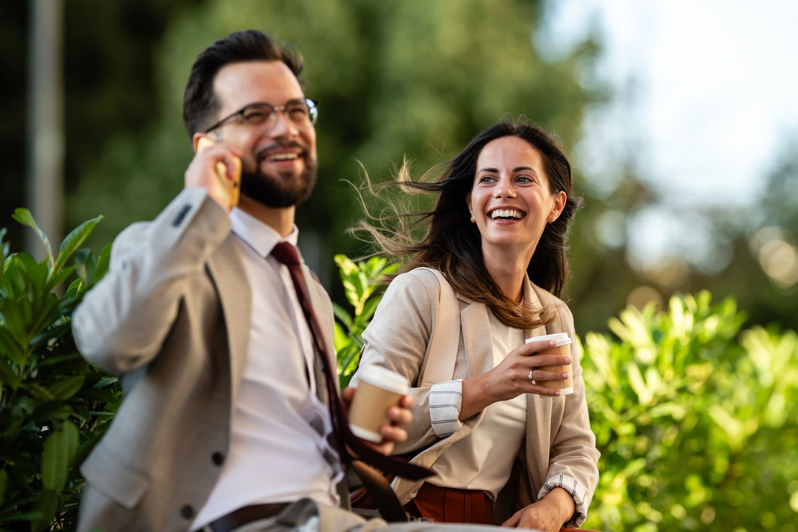 Two colleagues enjoying coffee outdoors, one on the phone.