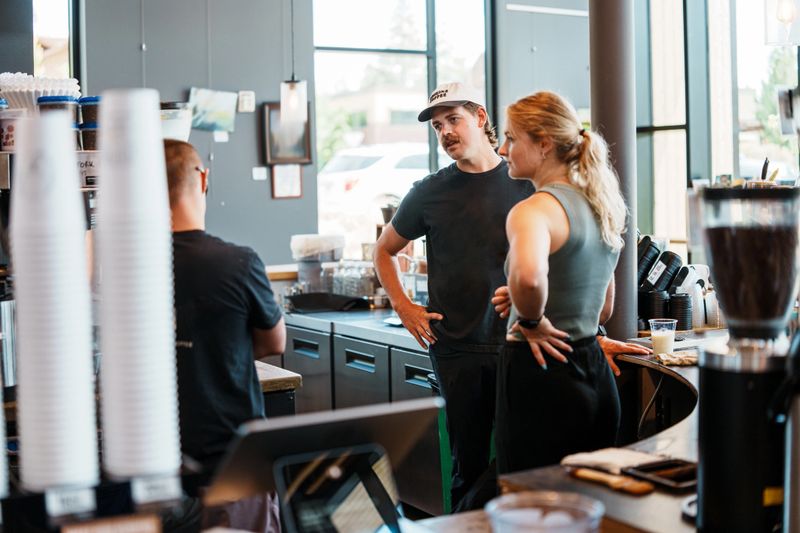 A coffee shop manager speaks with two team members behind the counter, fostering collaboration and hands-on leadership during a shift.