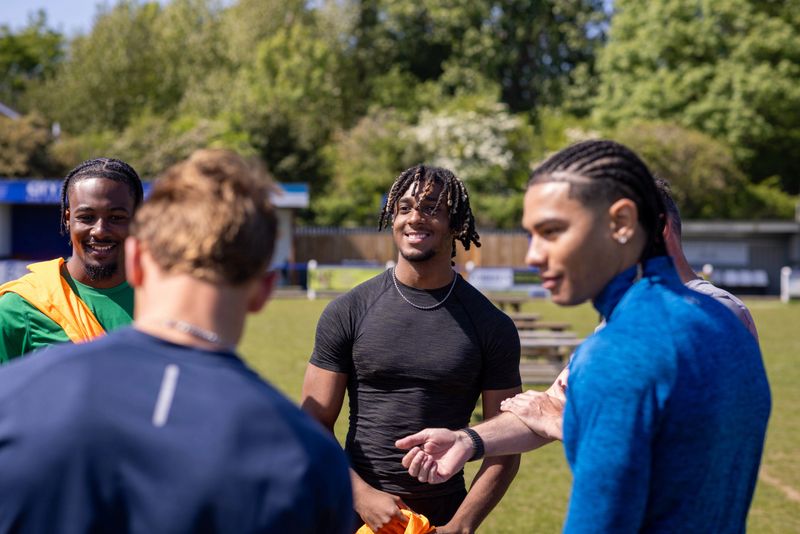 A medium view of a mature football coach talking to a diverse group of young male amateur footballers, they are standing around a table near the football pitch at football training in the North East of England. The coach is giving a pep talk before the team starts football practice. They are wearing sports clothing.Videos are available for this scenario.