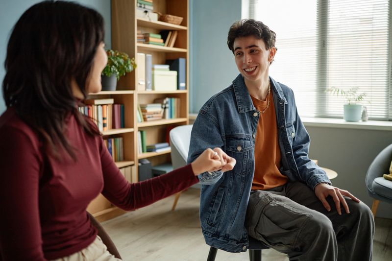 Caucasian teenage boy sitting in counseling session smiling and fist bumping with female counselor, bookshelf and window in background, engaging in supportive conversation