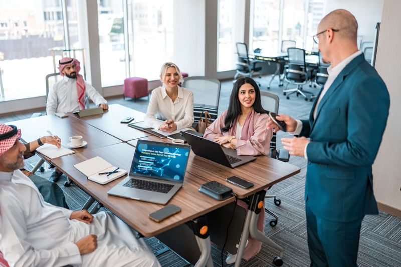 A mid adult Caucasian businessman in a blue suit leads a productive strategy meeting with a diverse team in a modern, well-lit office space.