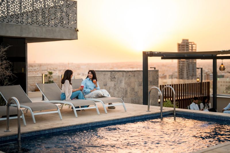 Lifestyle portrait of two young Middle Eastern women in casual resort wear conversing on white loungers beside a luxurious rooftop pool, city skyline glowing in warm sunset light.