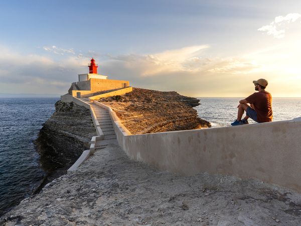 A man sitting near a lighthouse overlooking the ocean at sunset.