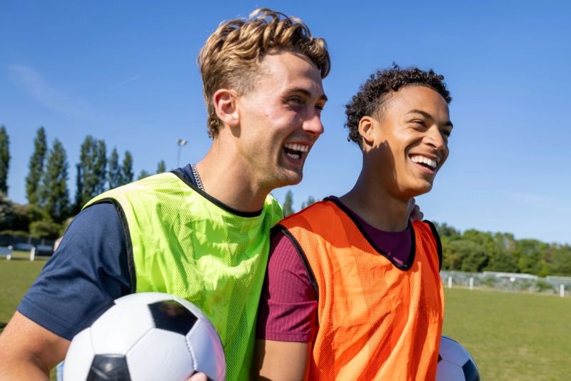 Young male football team leaving the pitch together after a match, talking and laughing with their arms around each other. They are at a football training ground in the North East of England. They're holding soccer balls, wearing sports bibs.