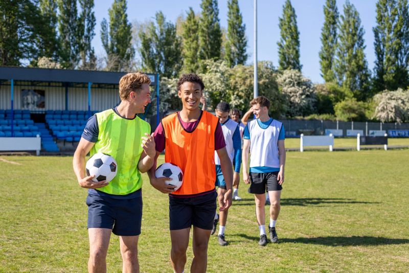 Young male football team leaving the pitch together after a match, talking and laughing with their arms around each other. They are at a football training ground in the North East of England. They're holding soccer balls, wearing sports bibs.