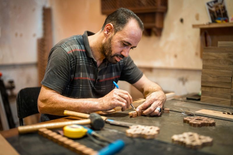 Close-up portrait of mid-adult Middle Eastern artisan wearing dark striped polo shirt, meticulously carving wooden mashrabiya patterns at workbench in traditional Al Balad workshop setting.