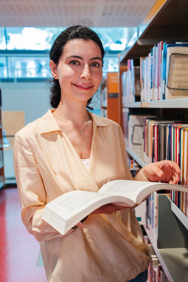Vertical. Charming Woman Smiling While Holding an Open Book in a University Campus Library Filled with Diverse Literature on Shelves, Creating a Cozy Intellectual Space for Reading and Learning. High quality photo