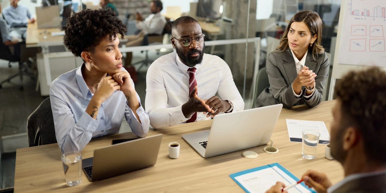 Four professionals engaged in a serious business meeting around a table with laptops.