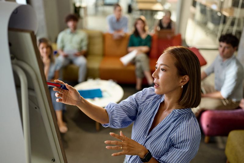 Korean businesswoman reading plans on whiteboard during sales pitch with her colleagues at casual office.