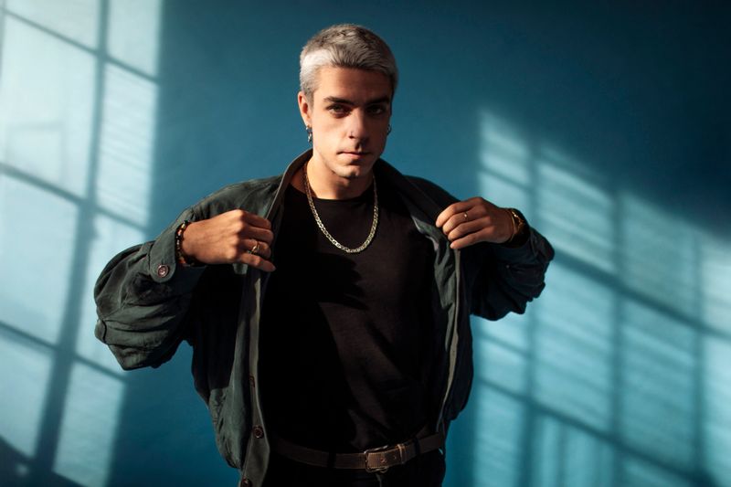 Portrait of a stylish young gen z man with short silver hair poses against the blue background under dramatic lighting, wearing a black shirt, jacket, and chain necklace and looking at the camera