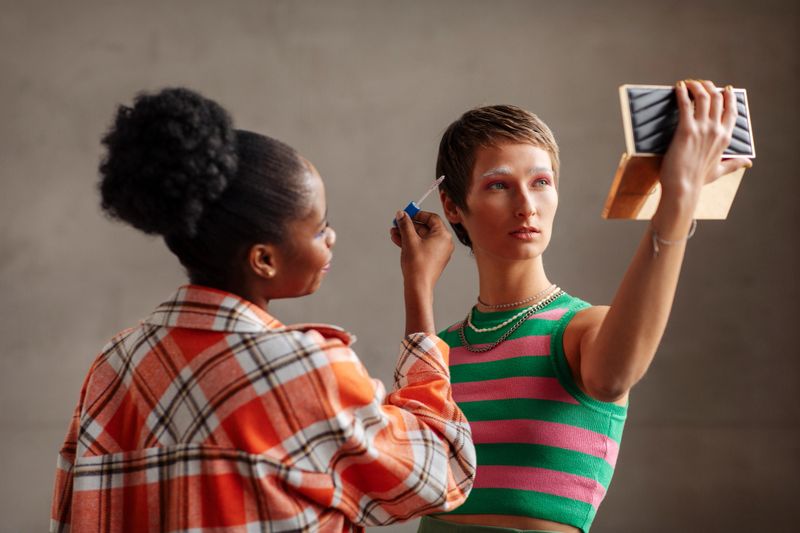 Two diverse Gen Z authentic girls indoors; One adjusts makeup while the other poses confidently, holding a mirror and looking at it