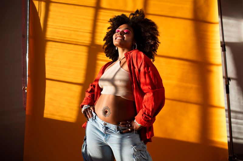 A young multicultural gen z girl confidently poses in a vibrant photography studio; She wears a red jacket and denim, illuminated by warm orange lighting