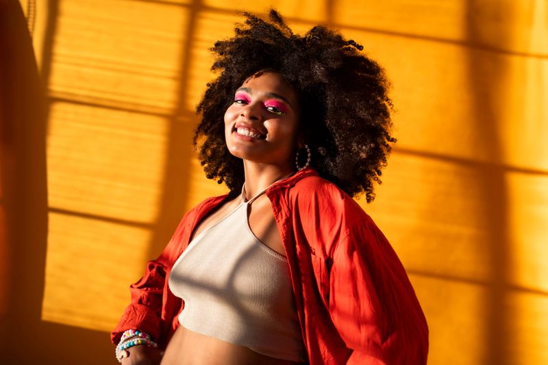 Portrait of a young multicultural gen z girl with vibrant makeup, natural curly hair, and a bold red jacket smiles confidently in a brightly lit photography studio
