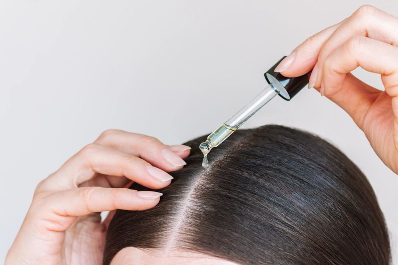 Close-up young brunette woman applying a drop of oil from pipette to her scalp. Hair loss. Strengthening and growth of hair.  Hair care.