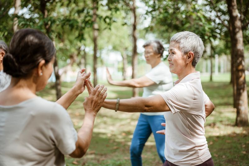 Older Asian people performing tai chi together in a calm, tree-filled outdoor space. Their gentle focus and posture reflect balance, strength, and the importance of companionship and routine in senior wellness.