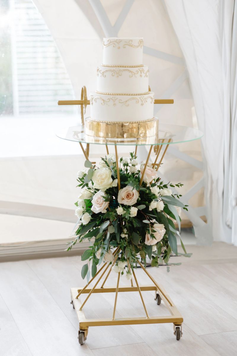 Delicious white wedding cake adorned with elegant gold decorations, resting on a glass stand and surrounded by beautiful floral arrangements