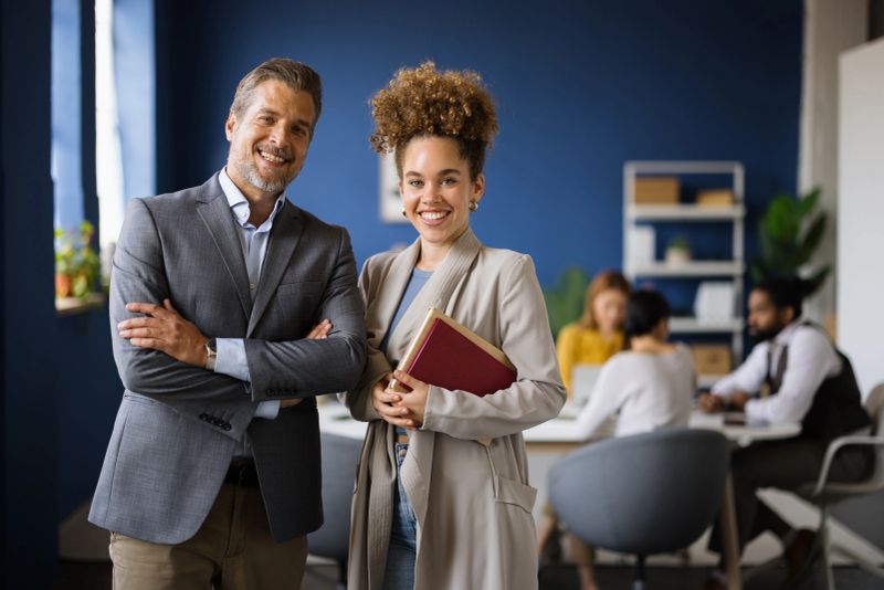 Portrait of two smiling business people standing with arms crossed in modern office with colleagues working in background