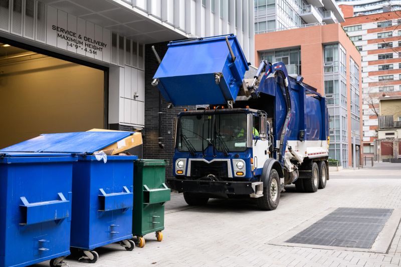 A front-loading garbage truck lifts a blue bin outside a city building's service entrance. Urban recycling and waste management in action.