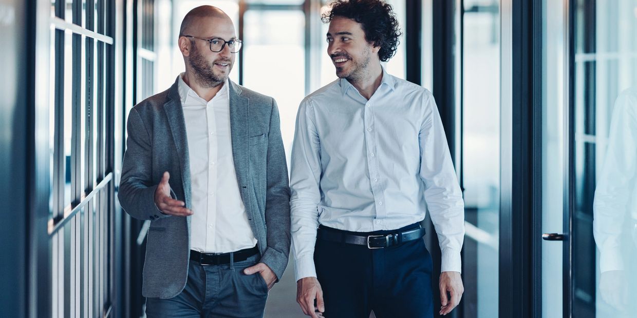 Two businessmen walking and discussing in a modern office corridor.
