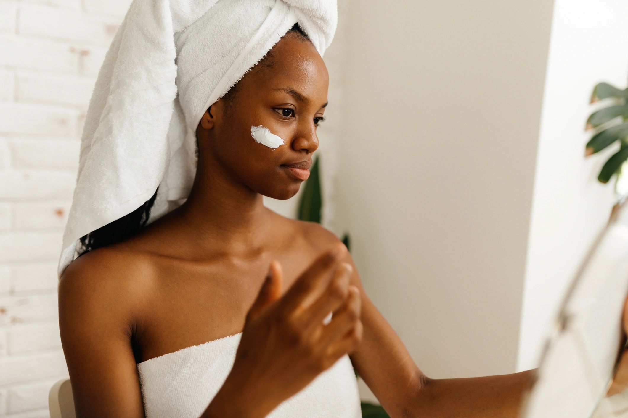 Woman with towel on head applying cream to her face in a bathroom.