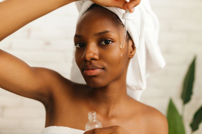 A young woman wrapped in a white towel with a towel on her head is applying a drop of facial serum on her cheek using a glass dropper. She is standing in front of a mirror with a calm and focused expression, surrounded by a minimal and bright setting that highlights her skincare routine and self-care moment.