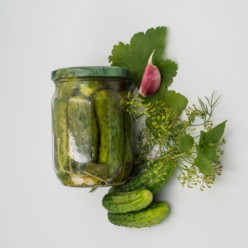 Стеклянная банка с маринованными огурцами, окружённая свежими огурцами, зубчиком чеснока, укропом и листьями петрушки на белом фоне. Идеально подходит для тем, связанных с ферментацией, заготовками, домашним питанием и здоровым образом жизни.