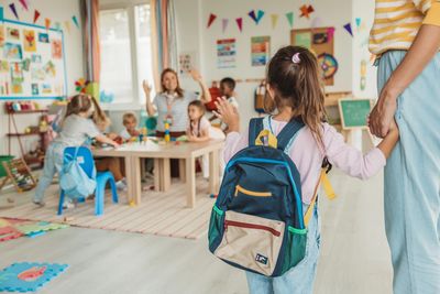 A young girl with a backpack holding an adult's hand in a colorful classroom.