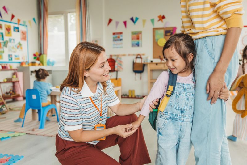 Preschool teacher in kindergarten classroom greeting new student, helping to easily assimilate with others.