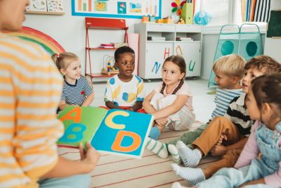 Children sitting in a circle learning ABCs from a teacher.