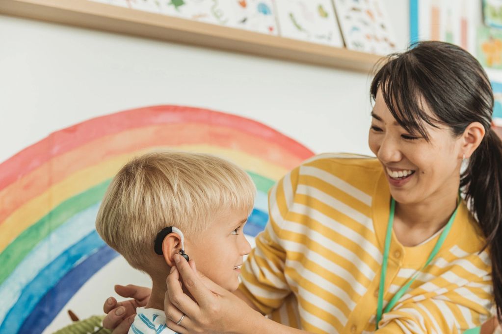Teacher helps child with hearing aid in colorful classroom.