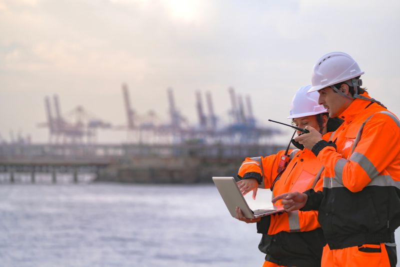Two engineers wearing safety uniforms and hard hats communicate using a radio while reviewing data on a laptop by the sea during sunset. Maritime industrial setting.