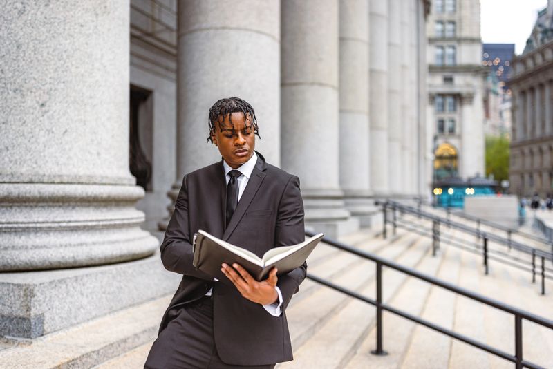 A young, professional man in a black suit stands outside a grand building with columns, reading a book, exuding confidence and focus in an urban environment.