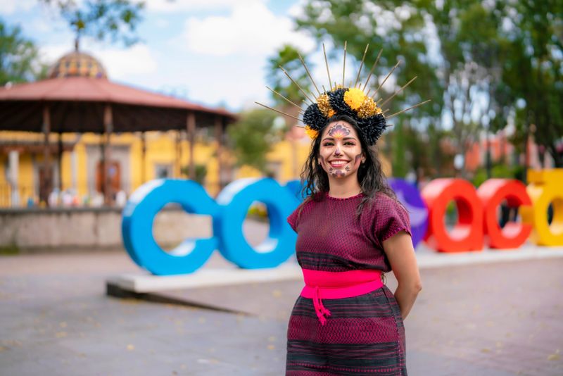 Young happy Latina colorful catrina makeup and handmade traditional dress smiling on a bench at Jardin Centenario in Coyoacan Mexico City vibrant look honors the spirit of Dia de Muertos with elegance