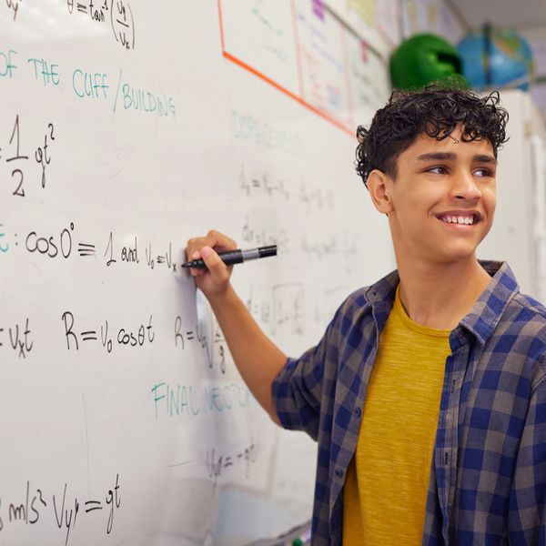 Student solving physics equations on a whiteboard, smiling.