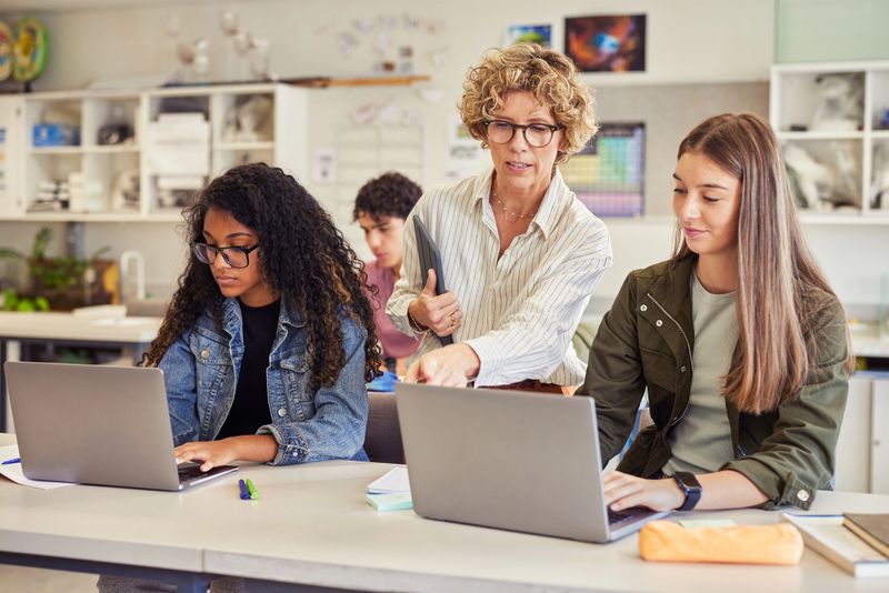 Mature teacher guiding student by pointing at laptop screen during computer class. Multiethnic students studying on laptop while listening to the teacher's instructions. Collaborative classmates using laptop while tutor helping and explaining new program.