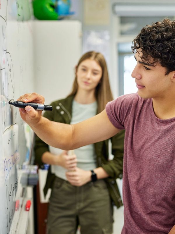 A young man writes on a whiteboard while a woman watches attentively.