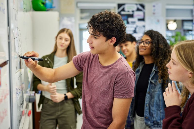 Male student writing on whiteboard while explaining a concept to his classmates at secondary school. Focused teens delivers a group presentation while drawing on board as classmates observe attentively. Teen guy helping classmates understand solution to geometry or arithmetic problem in classroom.