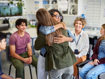 Two girls hugging while others watch in a classroom.