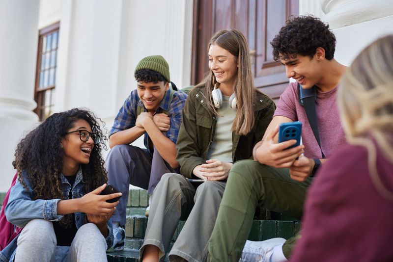Group of multiethnic students sitting on campus steps, smiling and sharing content on their smart phones. Happy guys and smiling girls having fun and watching video on cellphone. Group of young friends sitting together using phone app to share funny social media content.