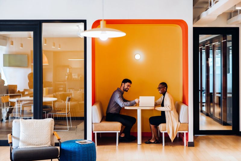 A man and woman engaged in a discussion while seated at a semi-enclosed office booth. Both are smiling and using a laptop, enhancing the collaborative atmosphere in this bright, vibrant space.