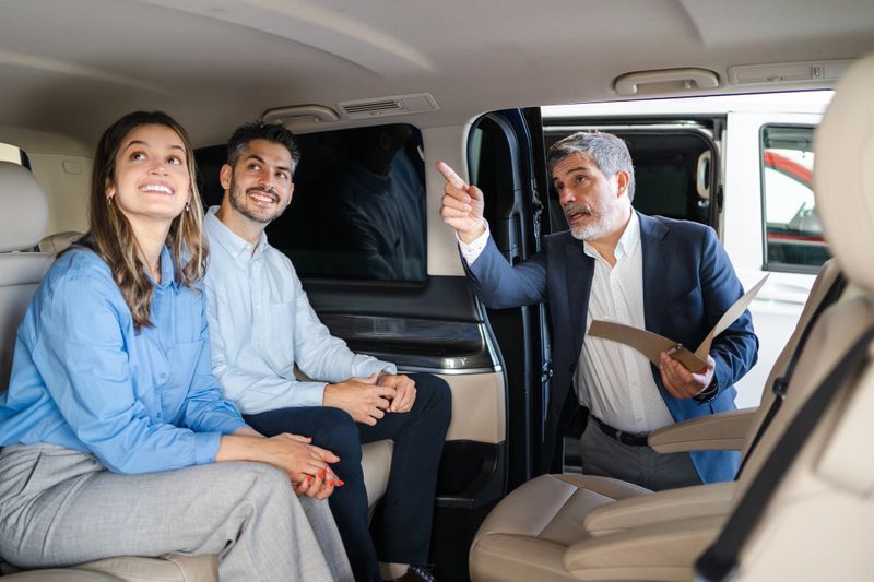 Car sales professional discussing vehicle features with young couple, comparing minivan capabilities and highlighting comfortable interior design