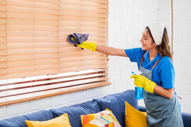 Smiling professional cleaner in blue uniform wipes window blinds in a modern living room. Highlights home cleaning services, sanitation, and attention to detail in residential housekeeping.