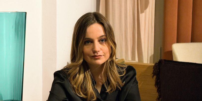 Woman with long hair in a black blouse sitting indoors.