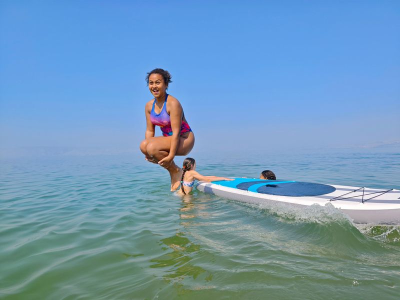 A smiling girl in a colorful swimsuit is captured mid-air in a tucked cannonball position, moments before splashing into the sea. In the background, friends lounge and swim around an inflatable paddleboard on a clear summer day. The bright blue sky, calm greenish water, and joyful expression combine to showcase the pure fun and freedom of youth, water play, and summertime adventure.