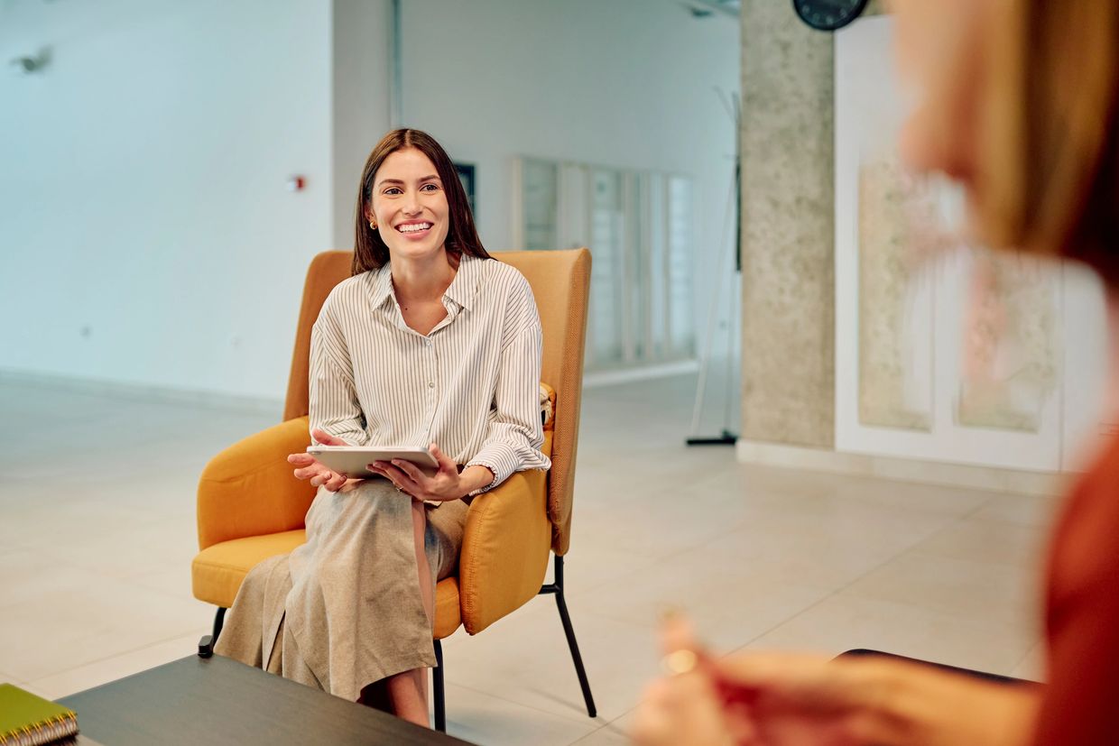Smiling woman sitting in a chair holding a tablet during a conversation.