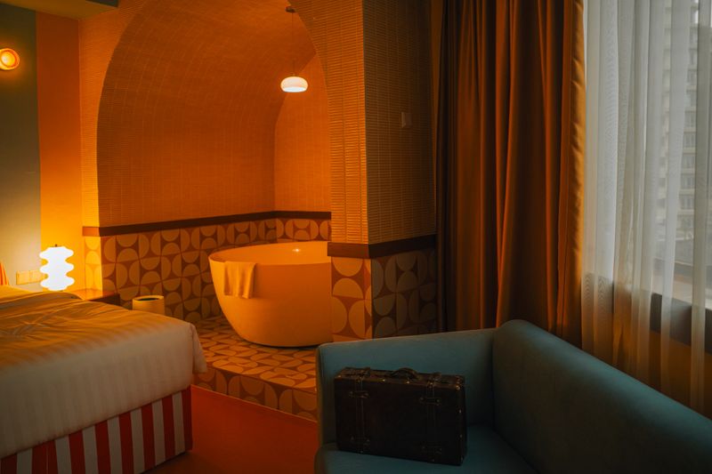 A warmly lit boutique hotel room featuring a retro-modern design with a freestanding round bathtub in a tiled alcove. The space includes a bed with striped base, vintage suitcase, soft blue armchair, and large curtained window letting in soft daylight. Captured in cinematic tones, evoking comfort and nostalgic charm. Perfect for travel, interior design, hospitality, or lifestyle themes.