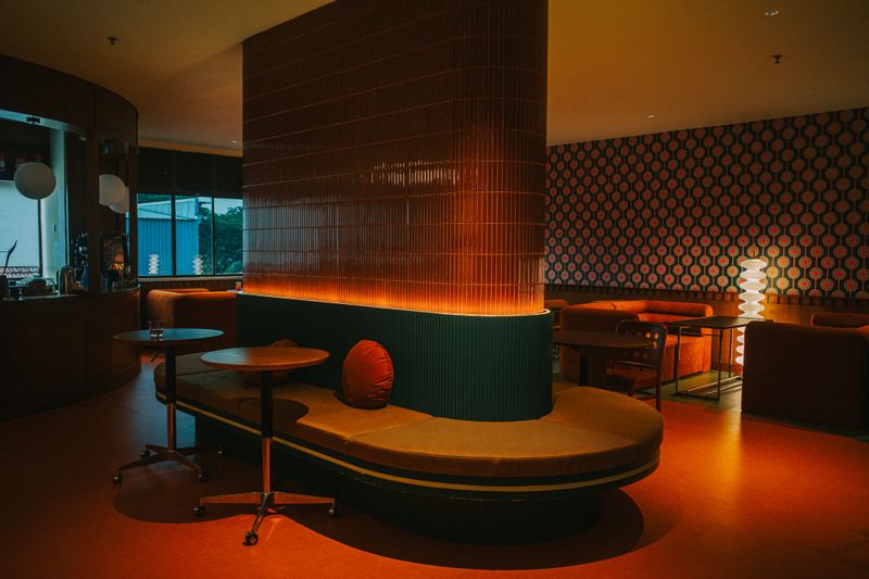 A cozy, retro-themed lounge interior featuring warm ambient lighting, mid-century modern furniture, and a curved tiled column with built-in seating. The space includes a coffee bar and a patterned feature wall, creating a relaxed and stylish atmosphere perfect for social gatherings or quiet moments.